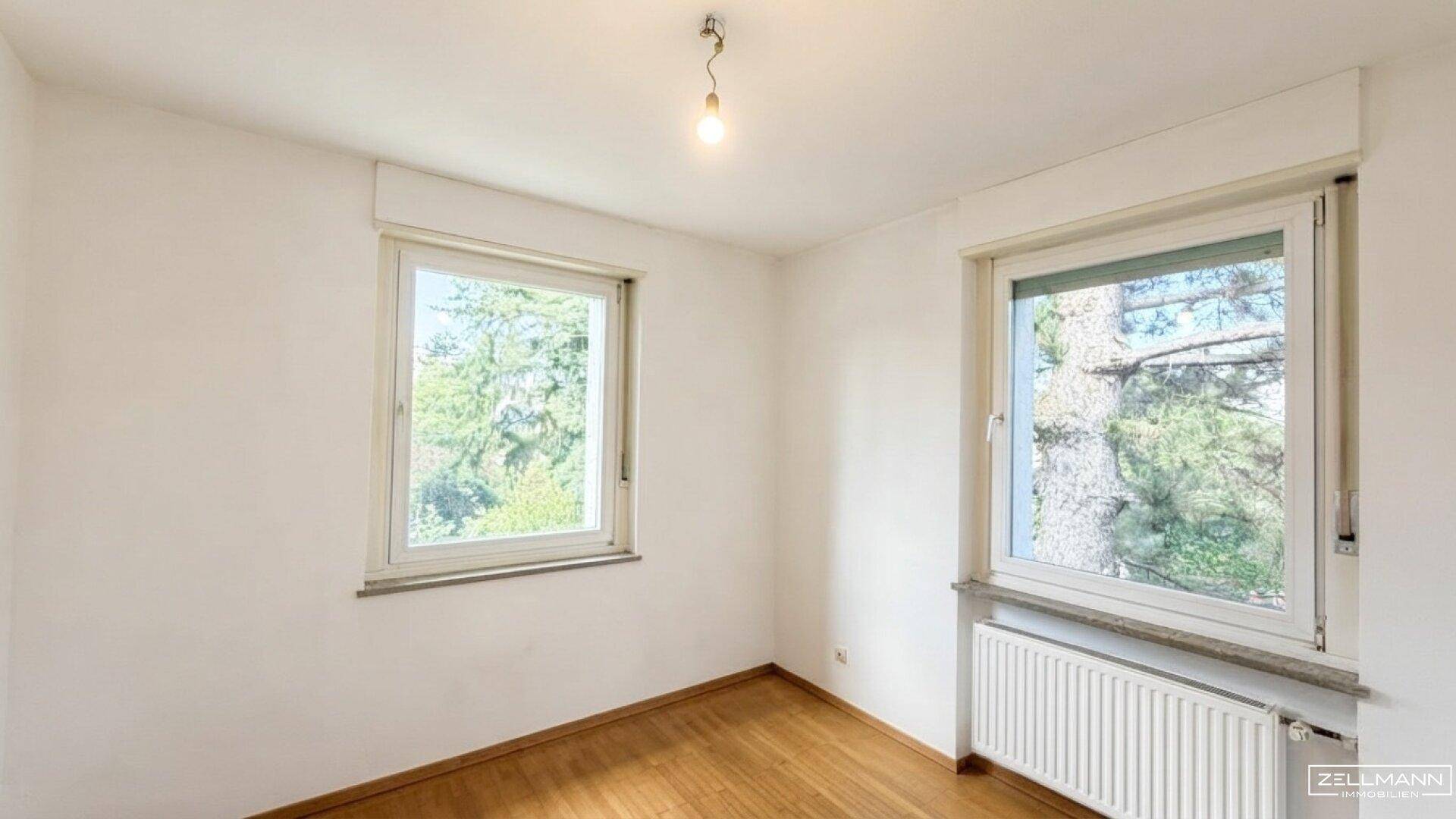 Helles Zimmer mit Holzboden und zwei Fenstern, die einen Blick ins Grüne bieten.