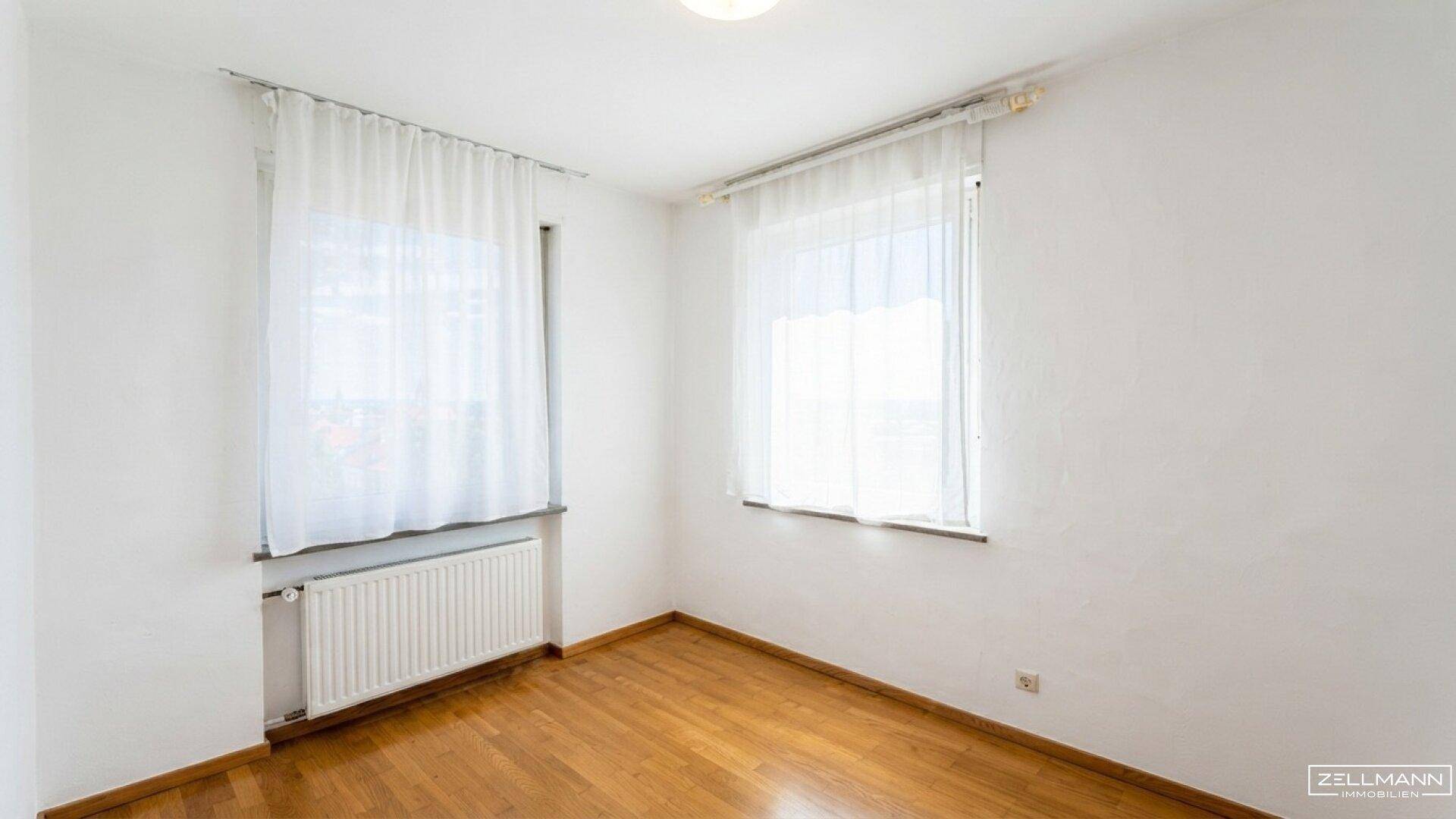 Helles Zimmer mit Holzboden und zwei Fenstern, die für ausreichend Tageslicht sorgen.