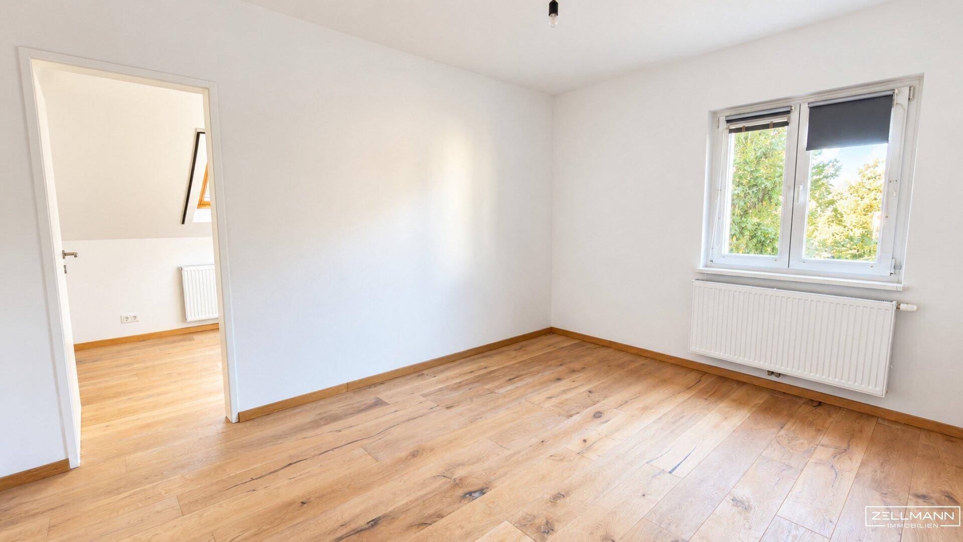 Leeres Schlafzimmer mit Holzboden, weiß gestrichenen Wänden und einem Fenster mit Tageslicht.