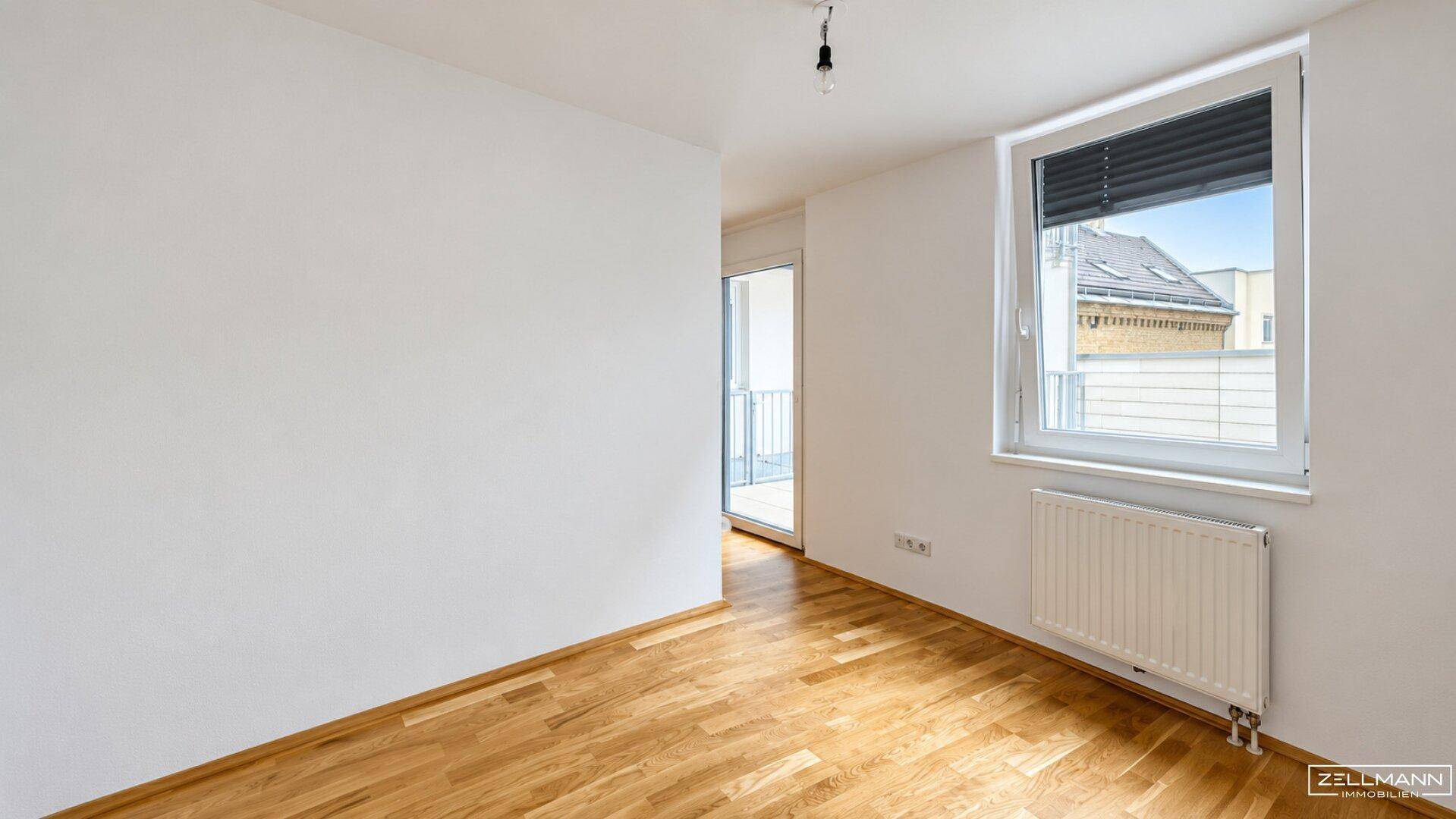 Helles Zimmer mit Holzparkettboden, großem Fenster und Heizkörper.