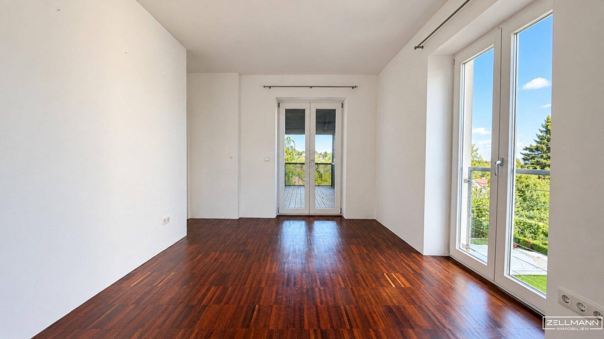 Heller Raum mit Holzboden und doppelten Fenstertüren, die ins Freie führen.