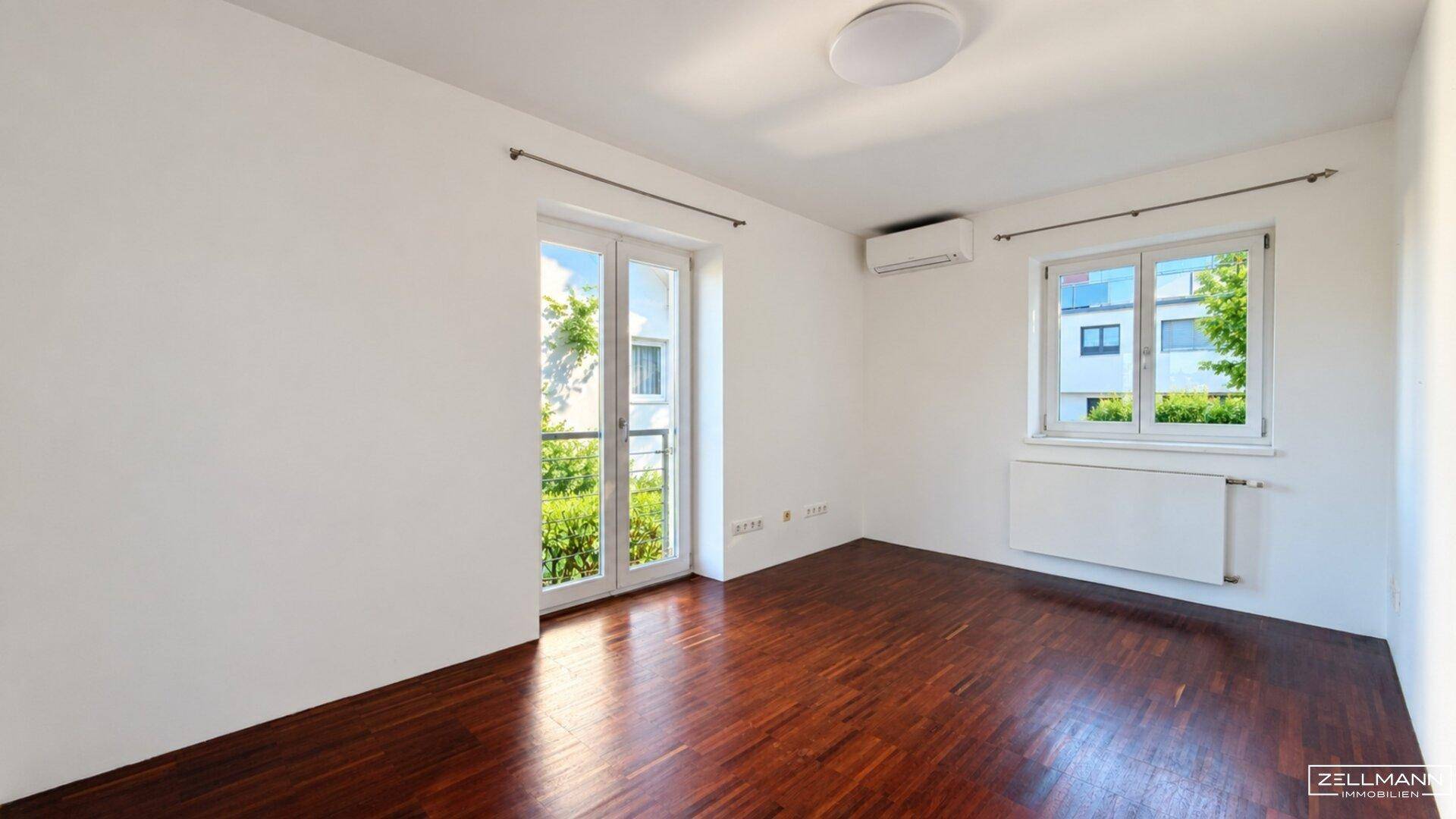 Geräumiges Zimmer mit dunklem Holzboden, großen Fenstern und Klimaanlage.