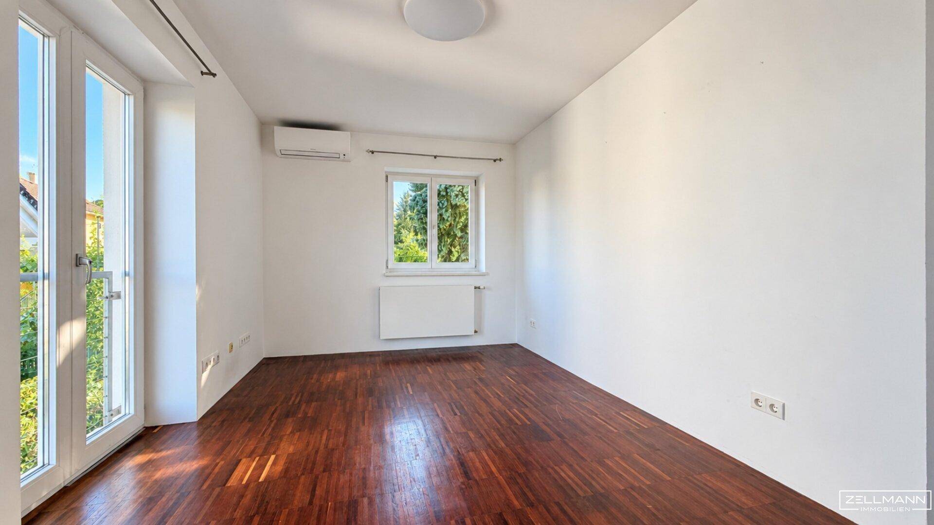Helles Zimmer mit Holzboden, Fenster und Zugang zu einem Balkon.