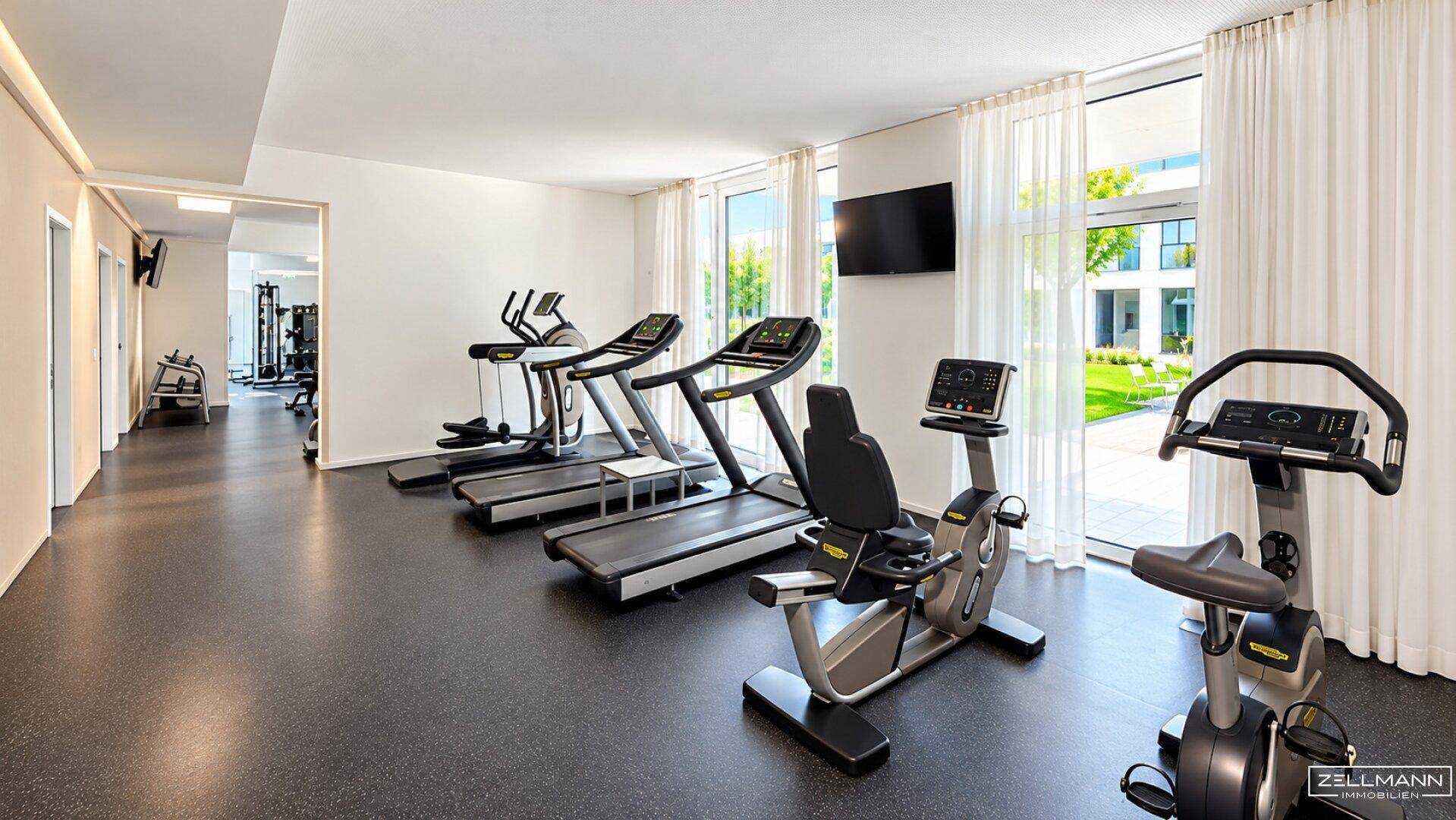 Moderner Fitnessraum mit Laufbändern und Fahrrädern, bietet viel Tageslicht durch große Fenster.