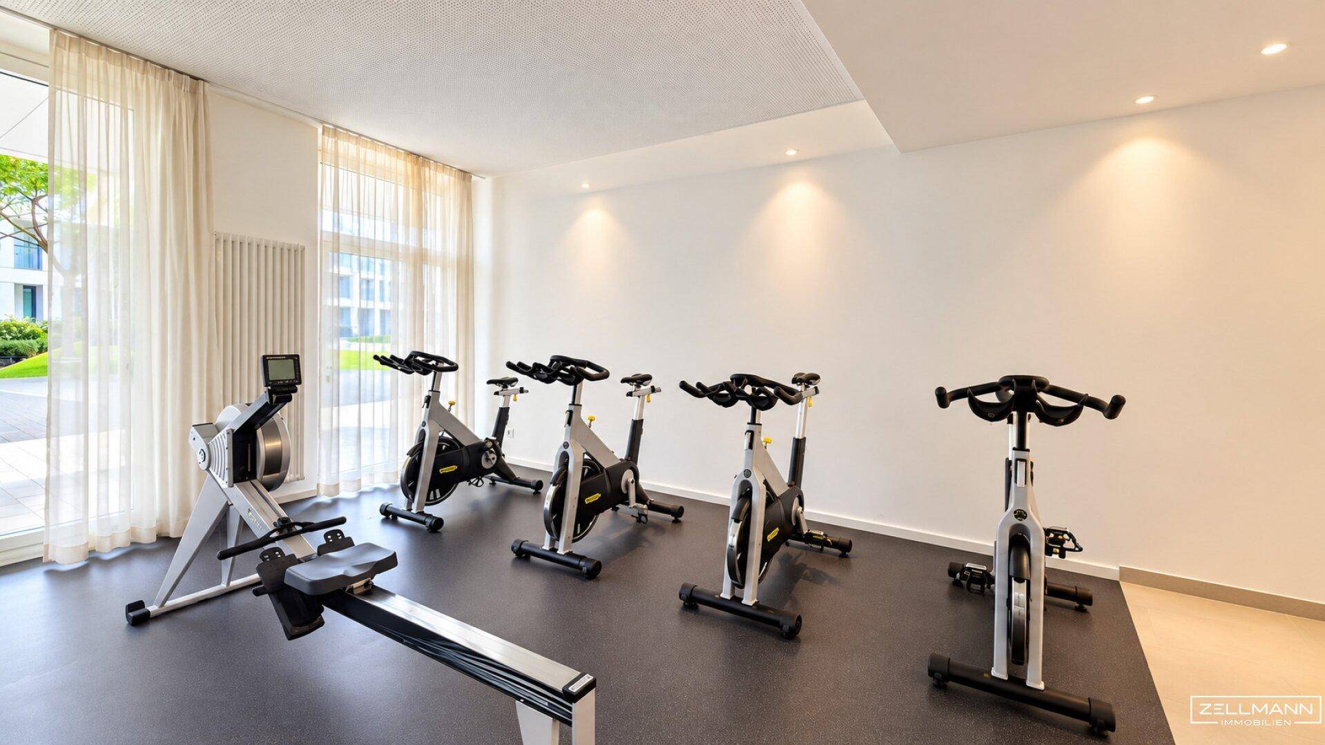 Weiterer Bereich des Fitnessraums mit Fahrrädern und Rudergerät, hell durch große Fenster.