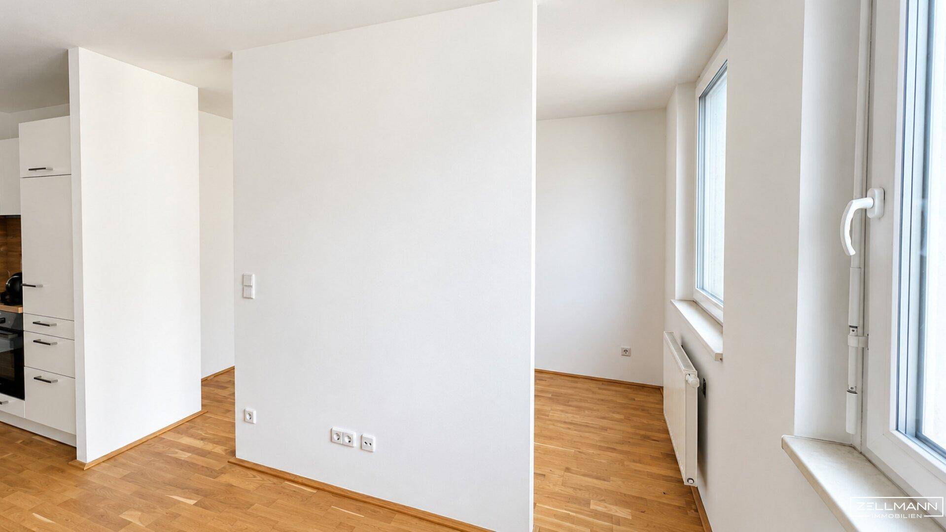 Lichtdurchfluteter Wohnbereich mit Holzboden und großen Fenstern, der an die Küche angrenzt.