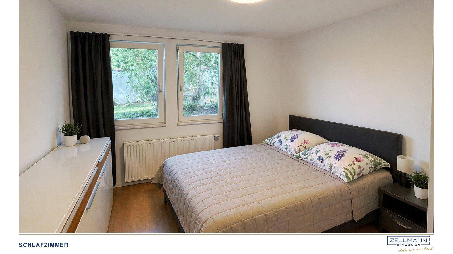 Schlafzimmer mit Doppelbett, Kommode und zwei Fenstern, die einen Blick ins Grüne bieten.