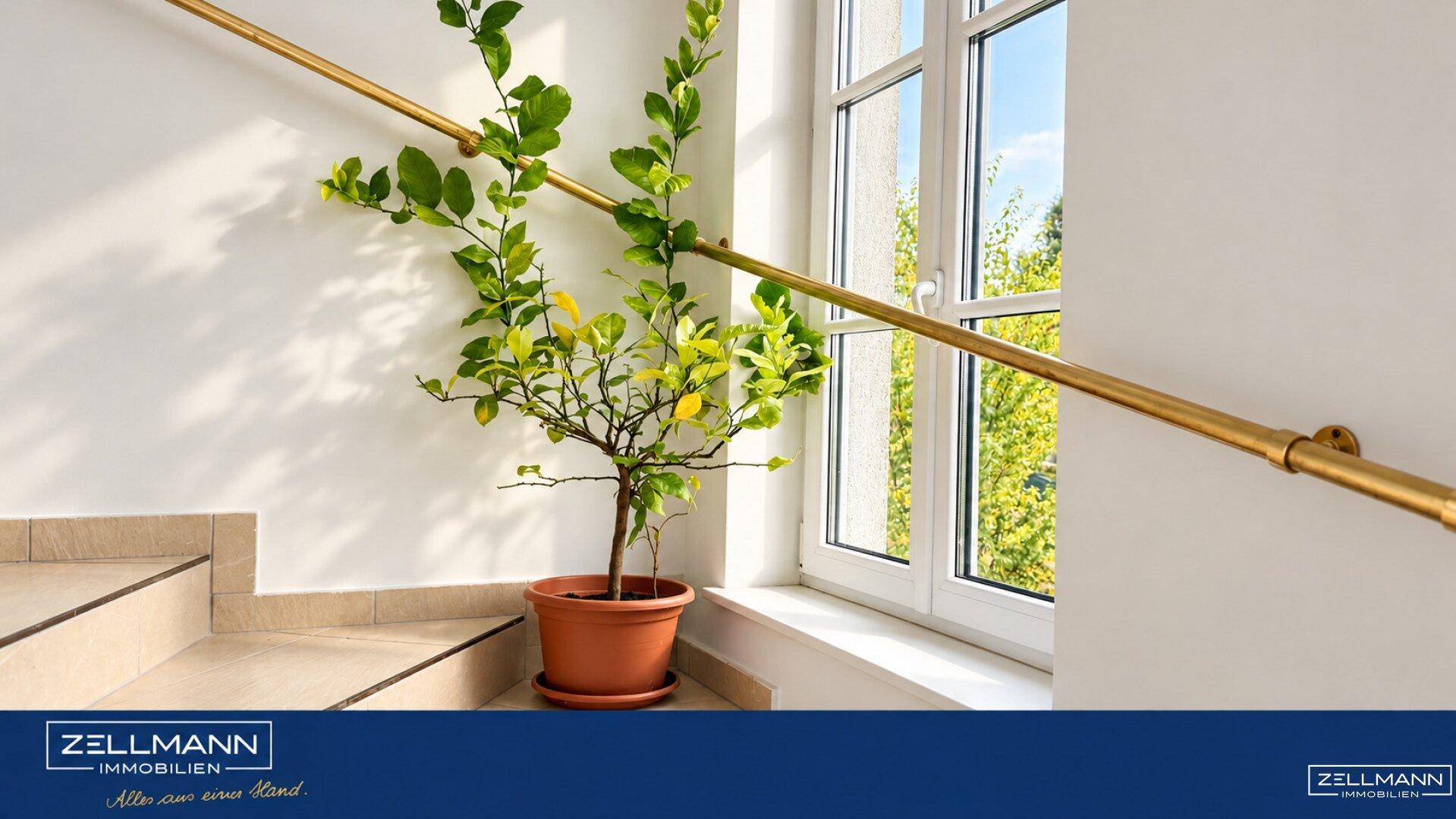 Treppenhaus mit hellem Fliesenboden, goldenem Handlauf und Fenster mit Blick ins Grüne.