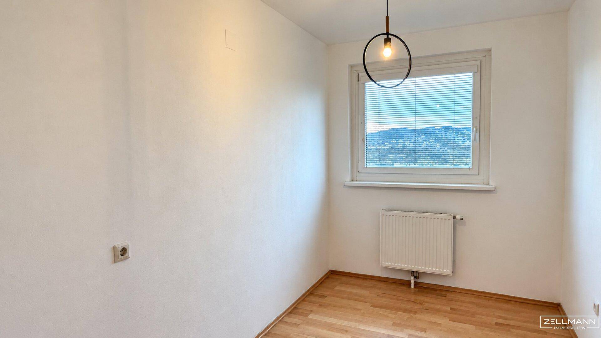 Kleineres, helles Zimmer mit Holzfußboden, weißen Wänden und einem Fenster mit Jalousien.