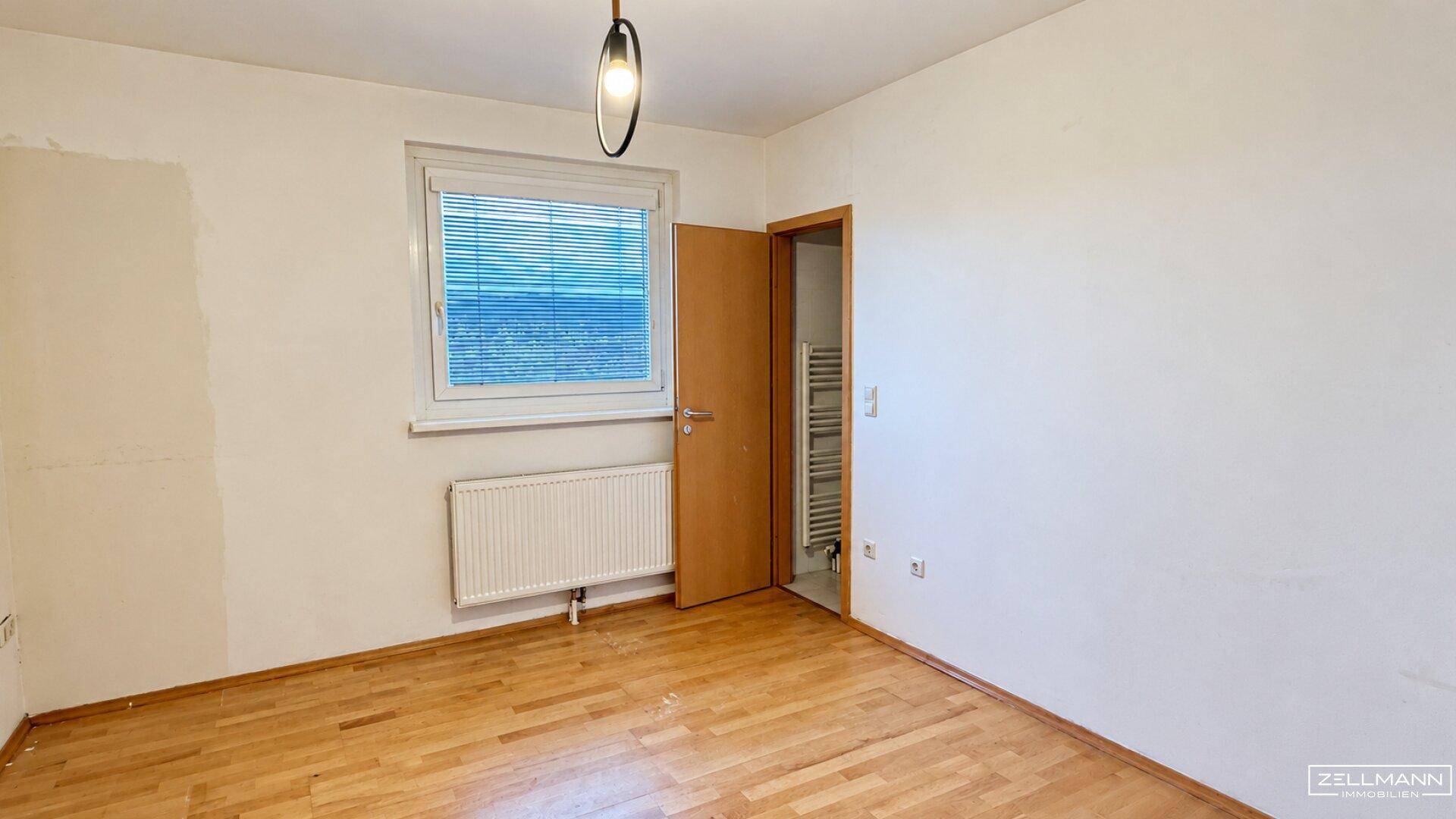 Leeres Zimmer mit Parkettboden, weißen Wänden und Zugang zu einem angrenzenden Badezimmer.