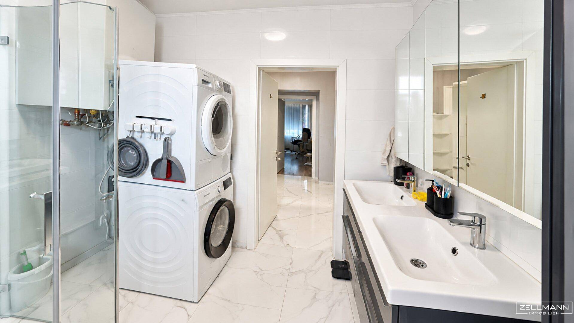 Helles Badezimmer mit Doppelwaschbecken, Dusche und gestapelter Waschmaschine und Trockner.