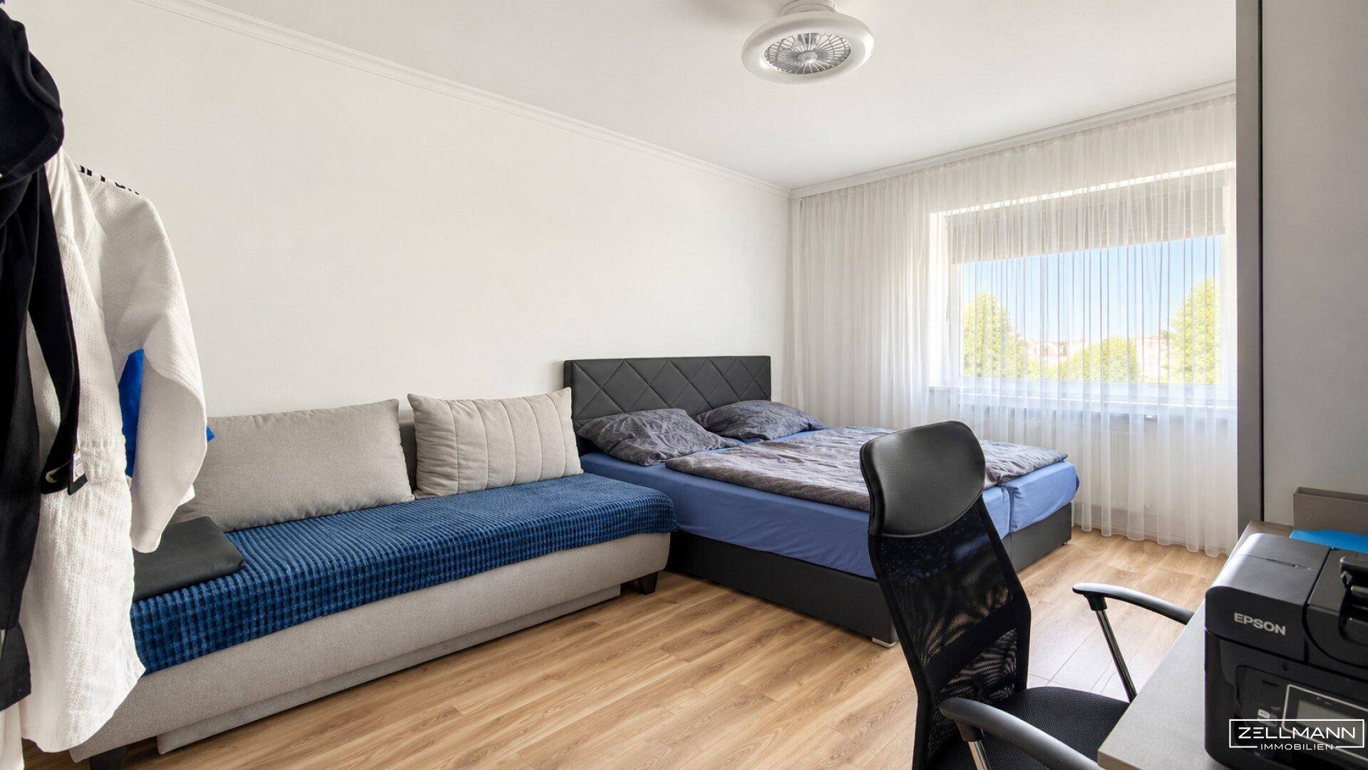 Schlafzimmer mit Bett, Sofa, Schreibtisch und großem Fenster mit Vorhängen.