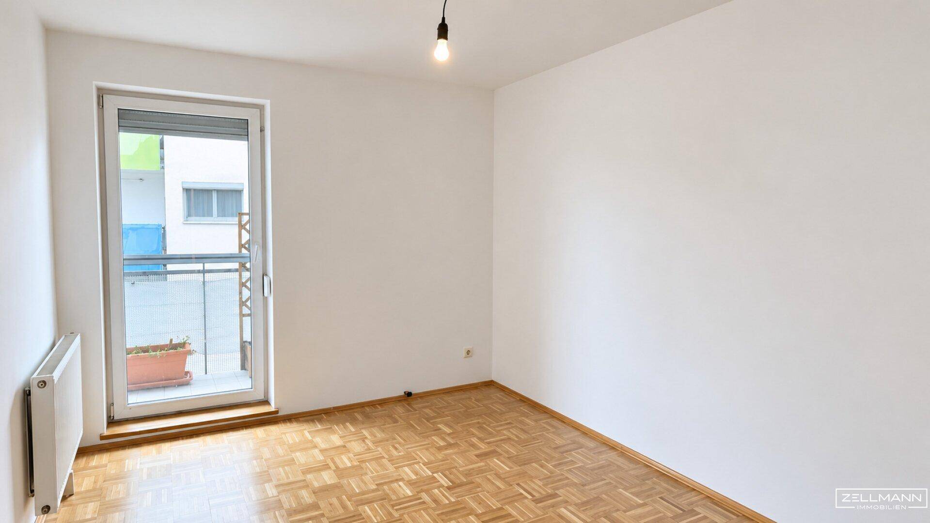 Helles, leeres Zimmer mit Parkettboden und Zugang zu einem kleinen Balkon.