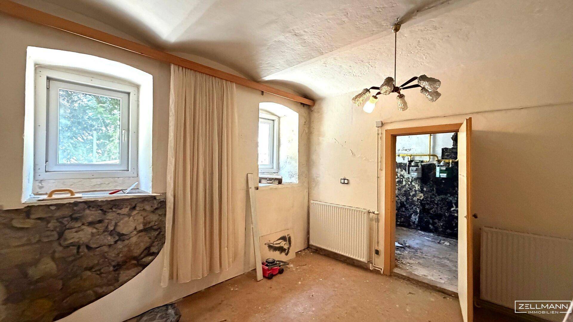 Renovierungsbedürftiger Raum mit freigelegten Steinwänden und zwei Fenstern, angrenzend an einen Technikraum.