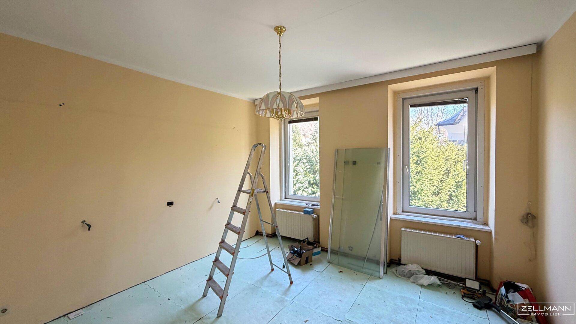 Geräumiger Raum mit zwei Fenstern und Renovierungsbedarf, ideal für einen Wohnbereich.