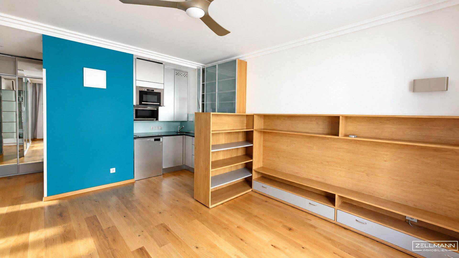 Heller Wohnbereich mit Holzboden, integrierter Küchenzeile und Deckenventilator.