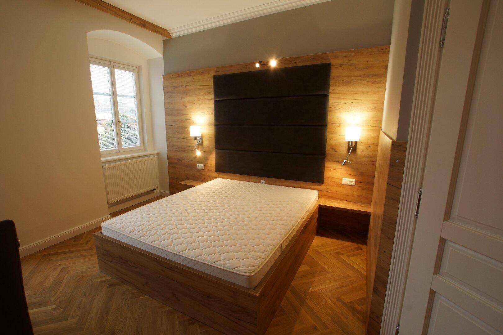 Schlafzimmer mit Holzbett, gepolstertem Kopfteil und Fenster mit Blick nach draußen.
