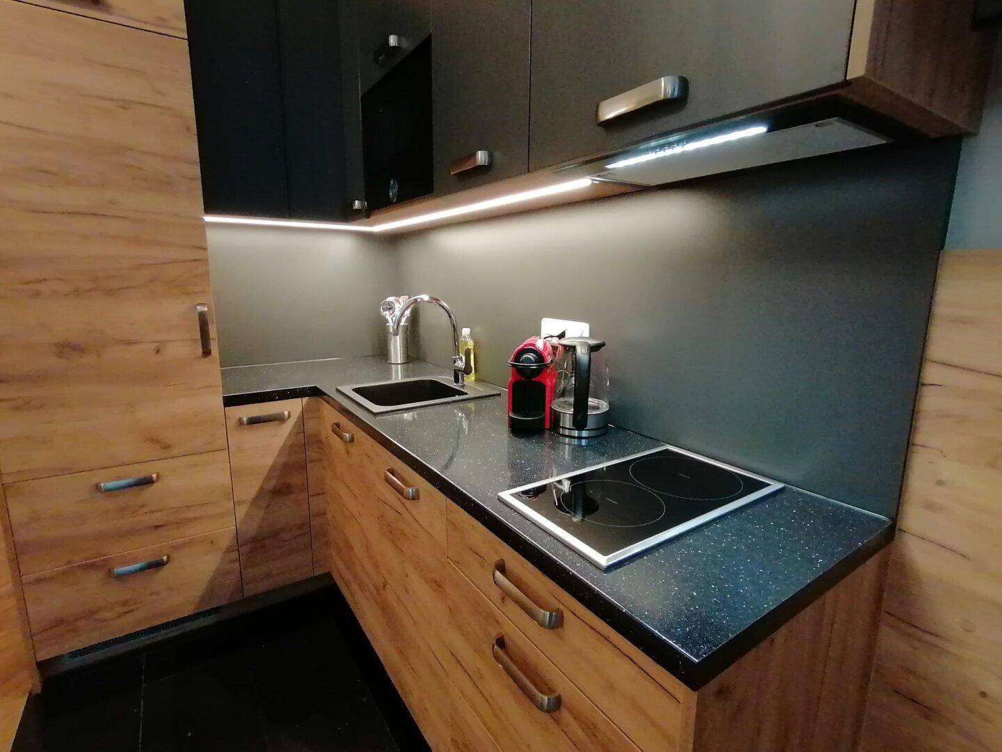 Moderne Küchenzeile mit Holzfronten, dunkler Arbeitsplatte, Induktionskochfeld und integrierter Beleuchtung.