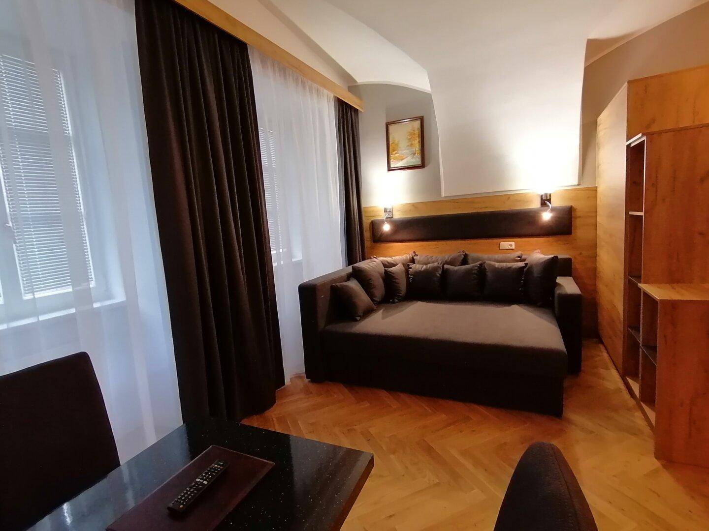 Gemütlicher Wohnbereich mit dunklem Sofa, Holzparkett und großen Fenstern mit schweren Vorhängen.