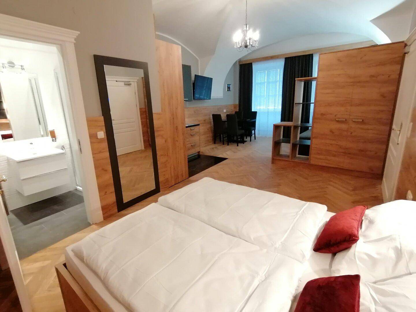 Schlafzimmer mit Doppelbett, Fischgrätparkett, großem Spiegel und Zugang zu einem modernen Badezimmer.