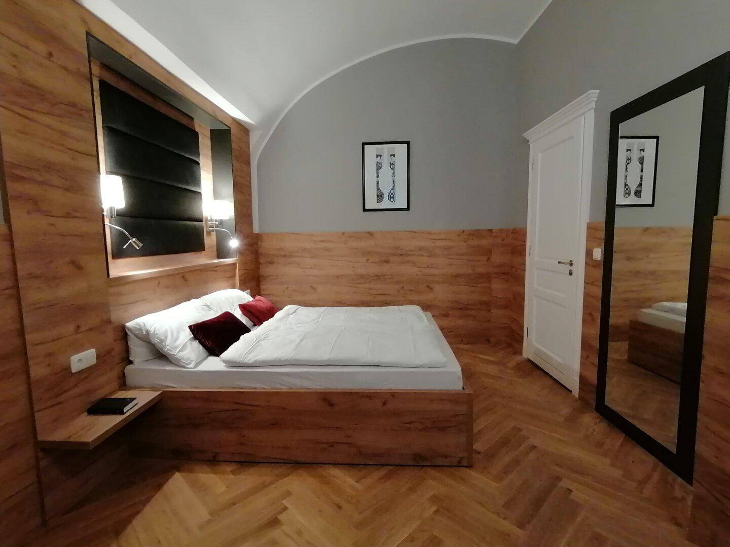 Schlafzimmer mit Holzbett, passendem Kopfteil, Fischgrätparkett und einem großen Wandspiegel.