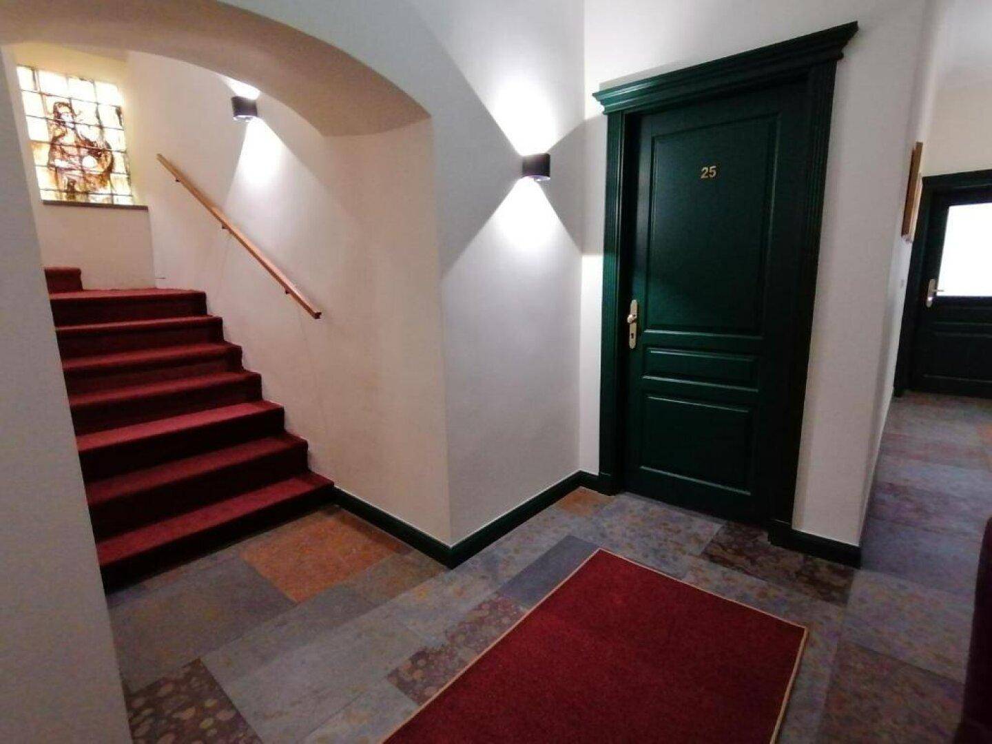 Treppenhaus mit rotem Teppich, dunkler Holztür und gemusterten Bodenfliesen sowie Buntglasfenster.