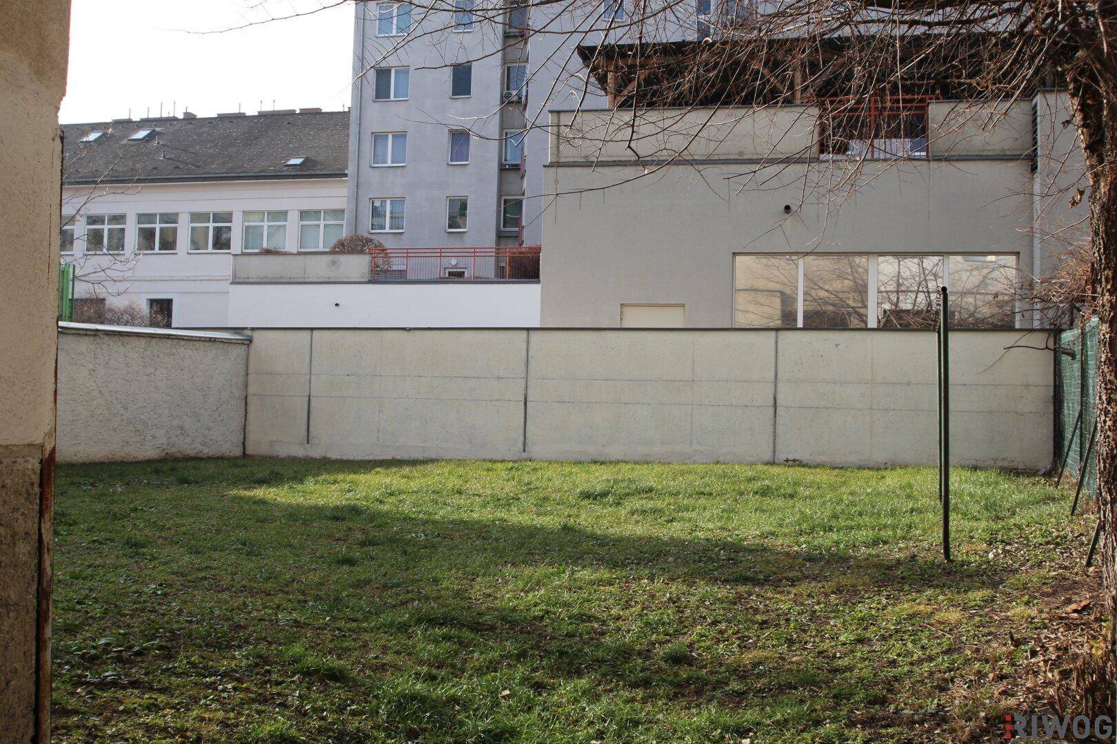 Grüner Gartenbereich mit Rasenfläche, umgeben von einer Mauer und Blick auf die Nachbargebäude.