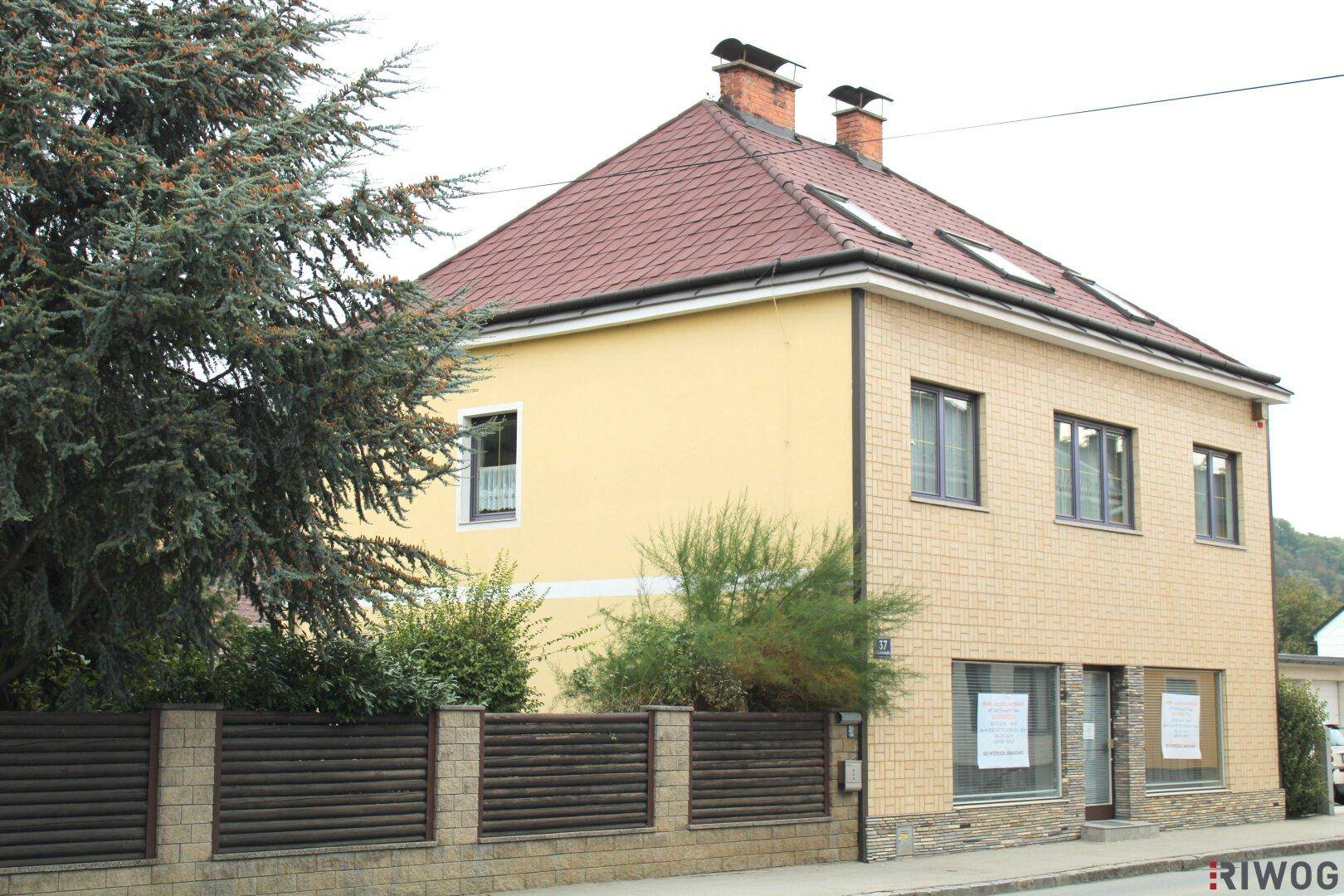 Gelbes Gebäude mit rotem Ziegeldach, teilweise verklinkerter Fassade und Schaufenstern im Erdgeschoss zur Straße.