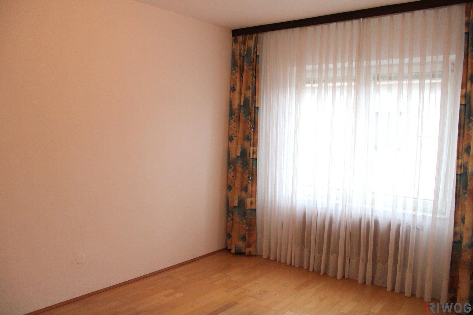 Heller Raum mit hellem Holzfußboden, einem Fenster mit Vorhängen und schlichten, hellen Wänden.