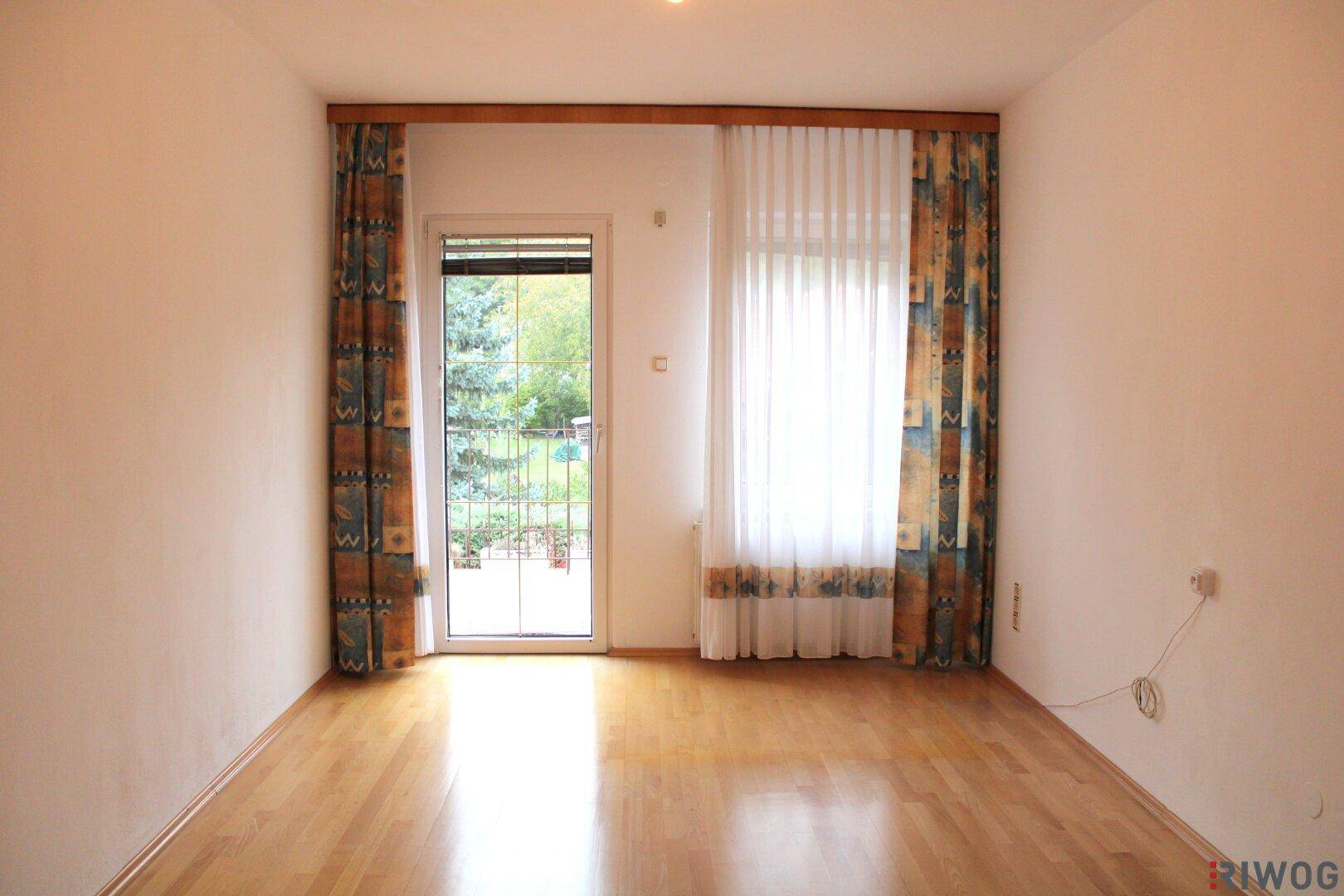 Leerer Raum mit hellem Holzfußboden, einer Tür zum Außenbereich und einem weiteren Fenster für viel Licht.
