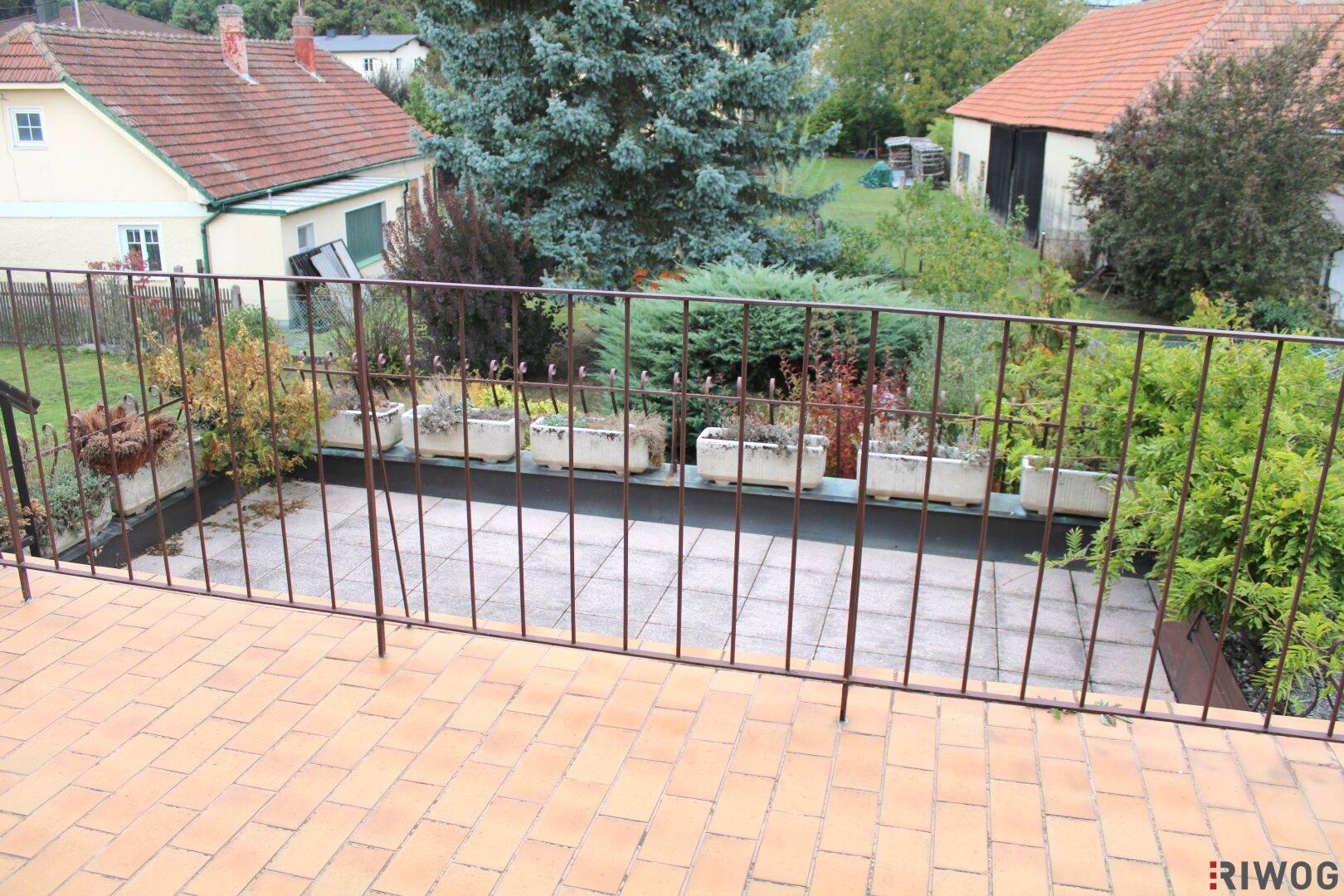 Blick von einem Balkon mit Fliesenboden und Metallgeländer auf eine gepflasterte Fläche und Grün.