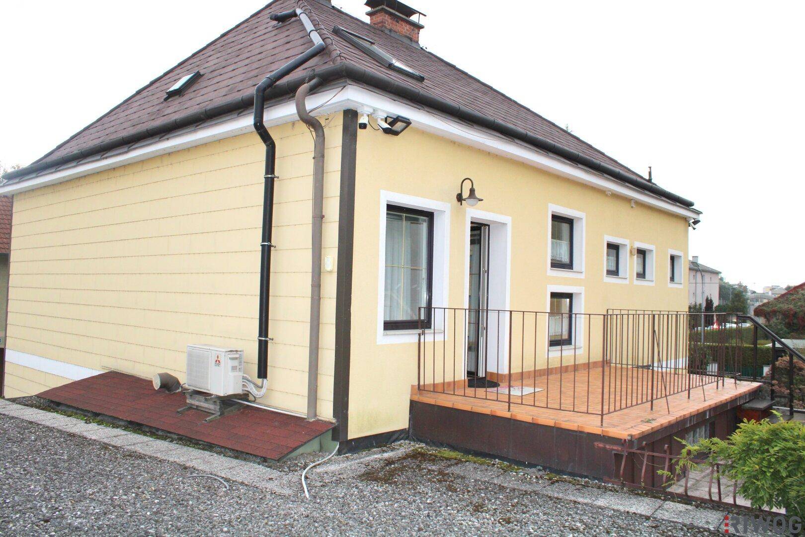 Seitlicher Blick auf das Haus mit kleiner Holzterrasse, Metallgeländer und einer Klimaanlage im Außenbereich.