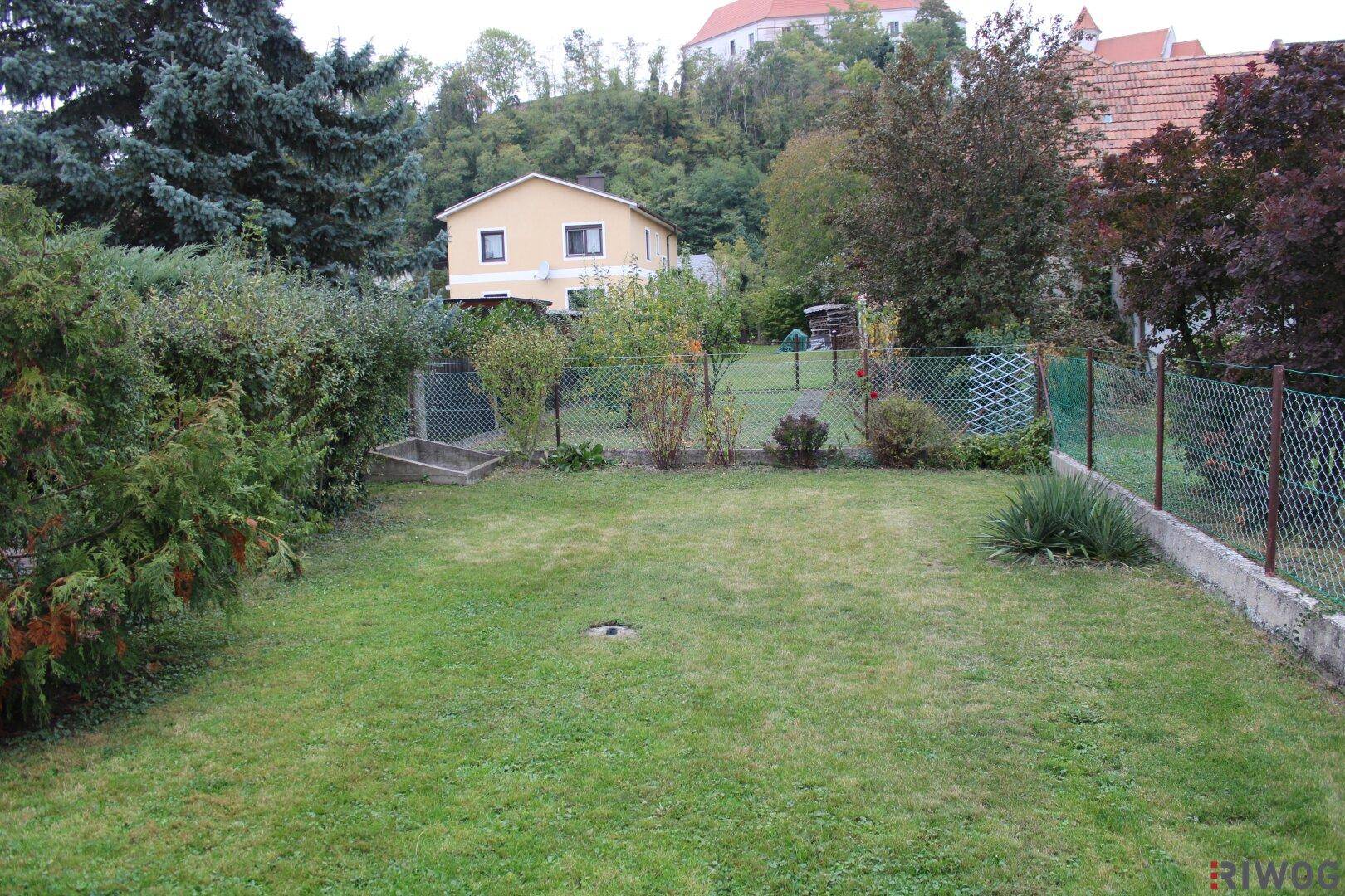 Hinterhof mit Rasenfläche, Zaun und einem entfernten Blick auf ein Gebäude auf einem bewaldeten Hügel.