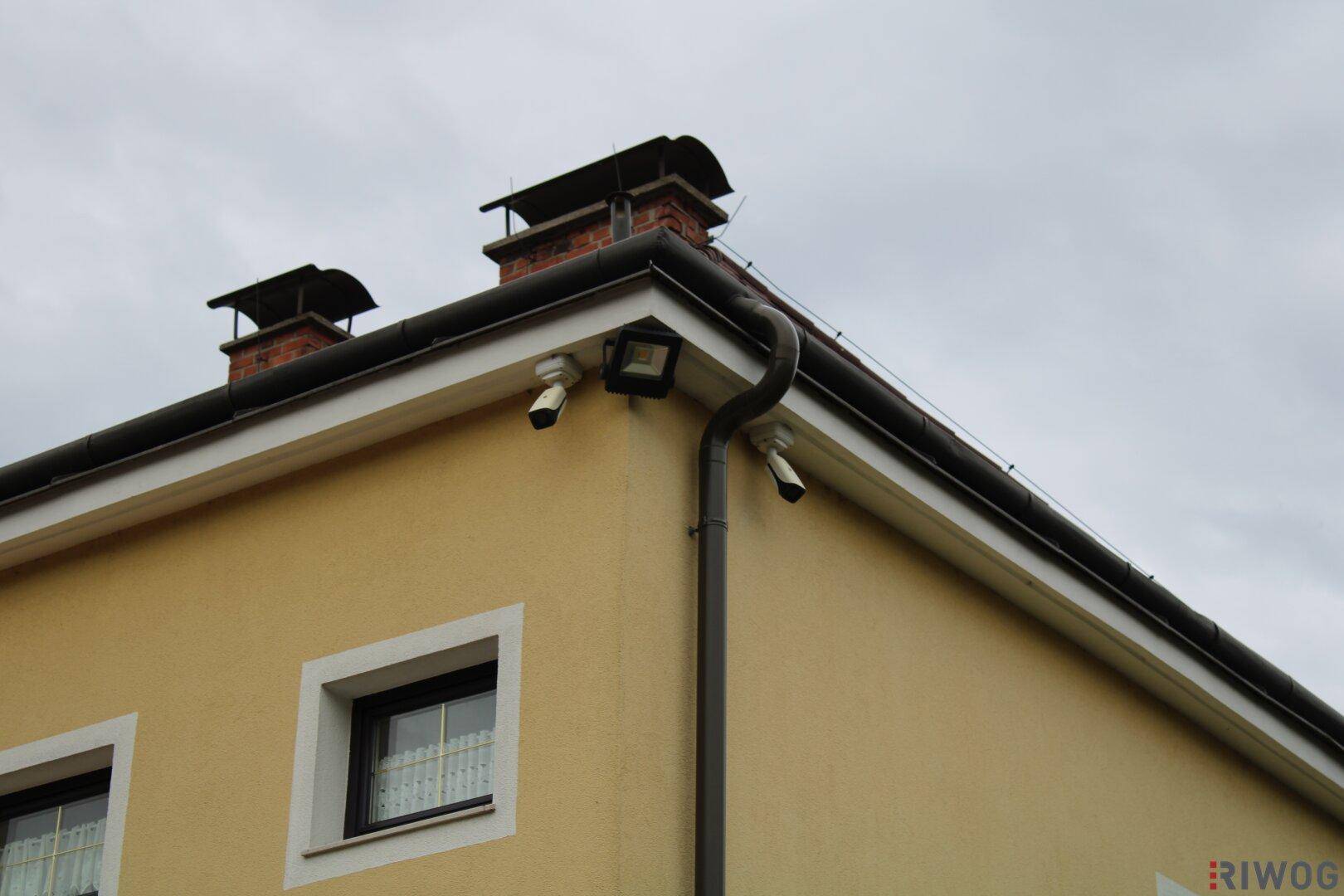 Nahaufnahme der Hausecke mit installierten Überwachungskameras und Außenbeleuchtung unter dem Dachgiebel.