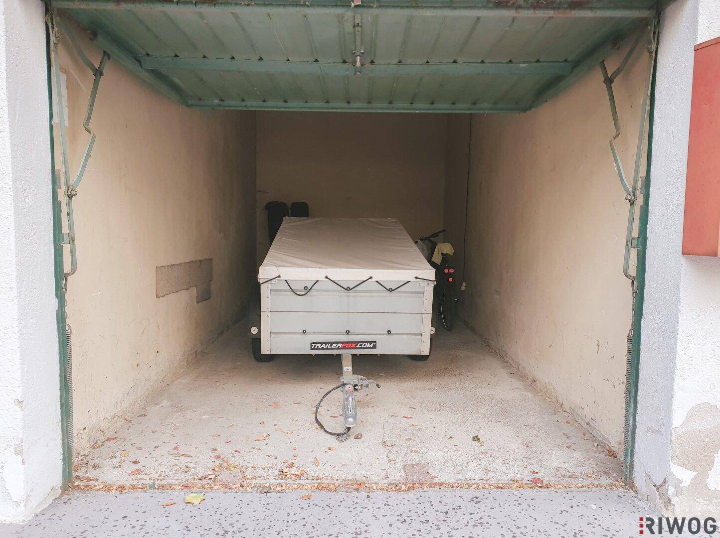 Innenansicht einer Einzelgarage mit geöffnetem Tor, einem Anhänger und einem Fahrrad auf Betonboden.