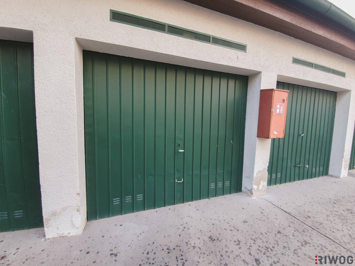 Reihe von Garagen mit grünen Metalltoren und einem roten Brandschutzkasten an der Fassade.