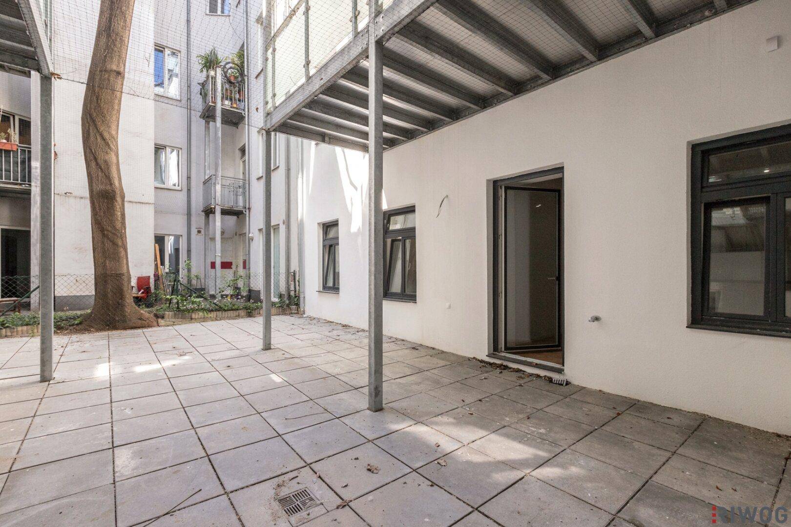Ruhiger Innenhof mit gepflastertem Bereich, einem Baum und überdachtem Zugang zu den Wohnungen.