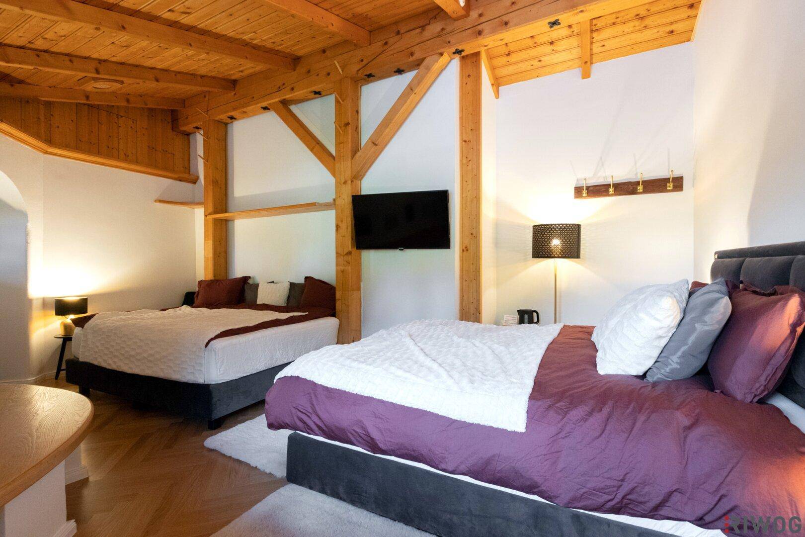 Geräumiges Schlafzimmer mit sichtbaren Holzbalken, zwei Betten und einem Fernseher an der Wand.