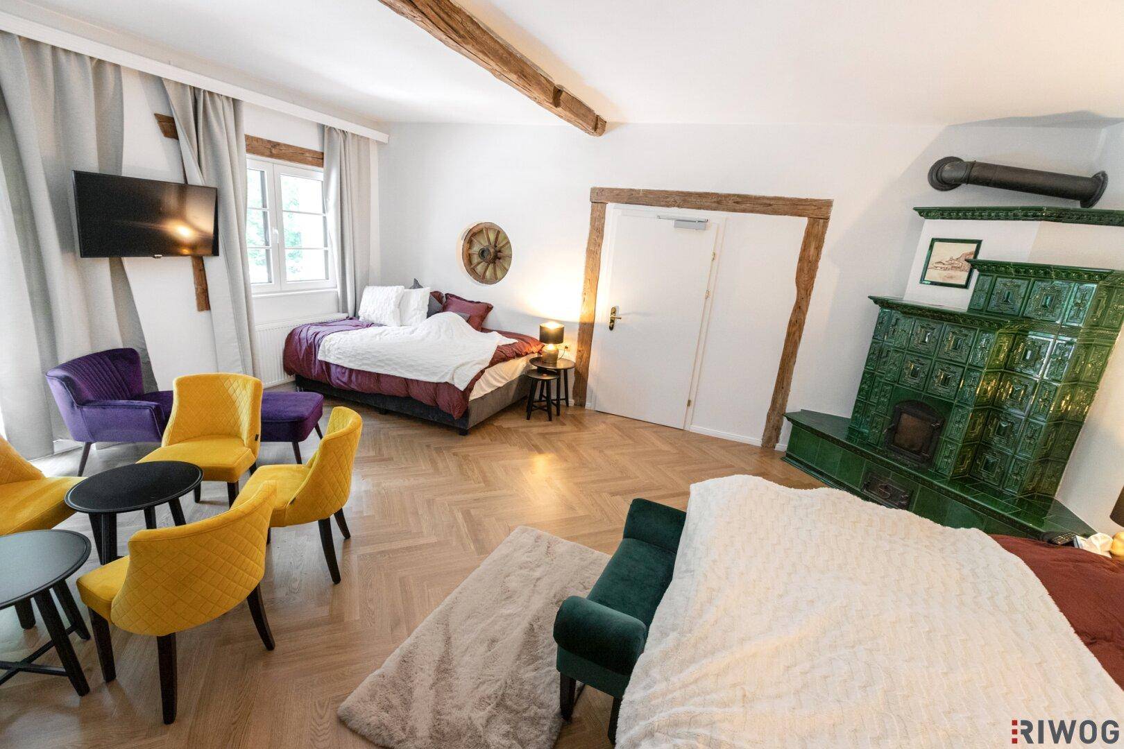 Geräumiges Schlafzimmer mit Kachelofen, Sitzgelegenheiten und Holzbalken an der Decke.