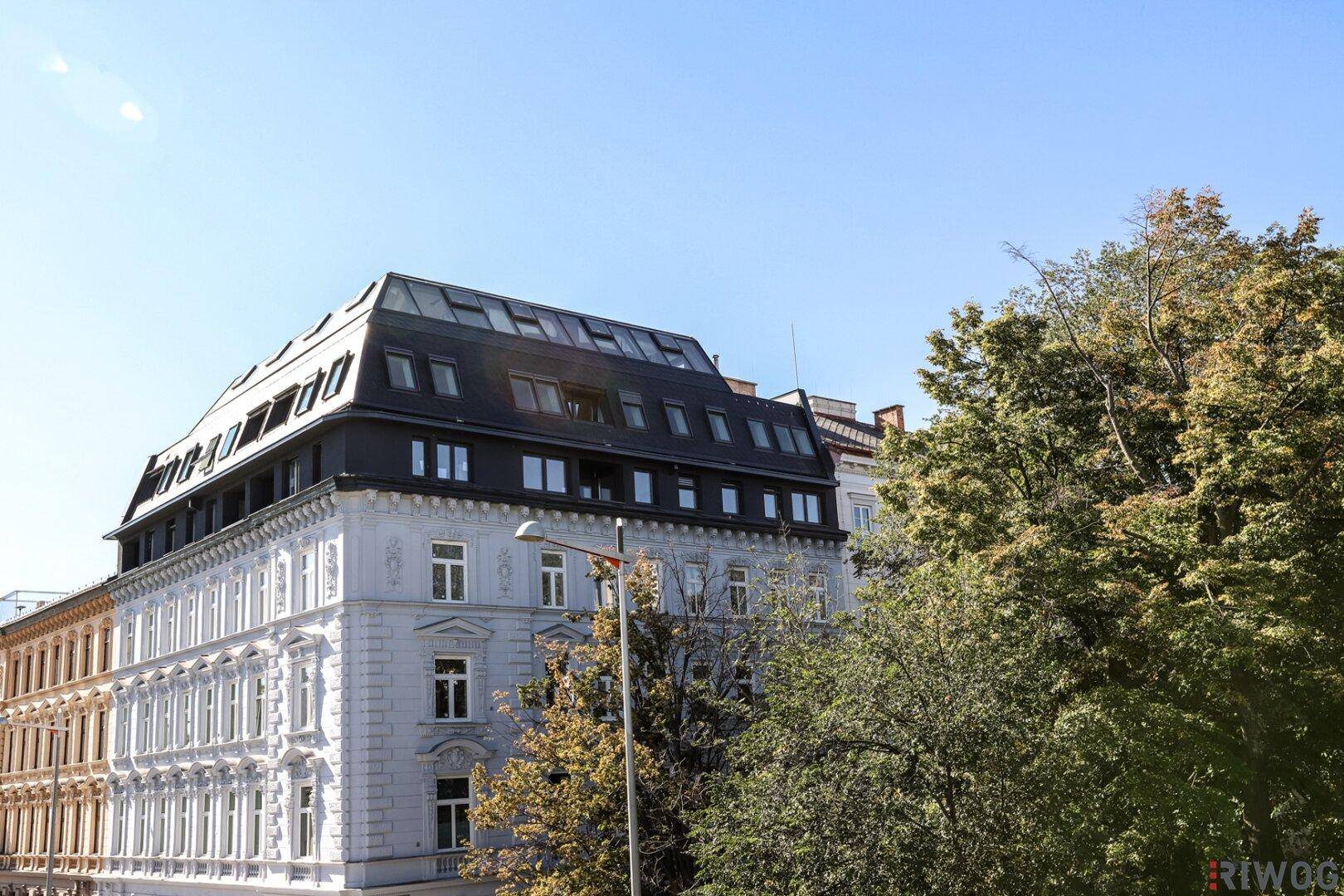 Gesamtansicht des sanierten Altbaus mit modernem Dachausbau und umgebenden Bäumen unter blauem Himmel.