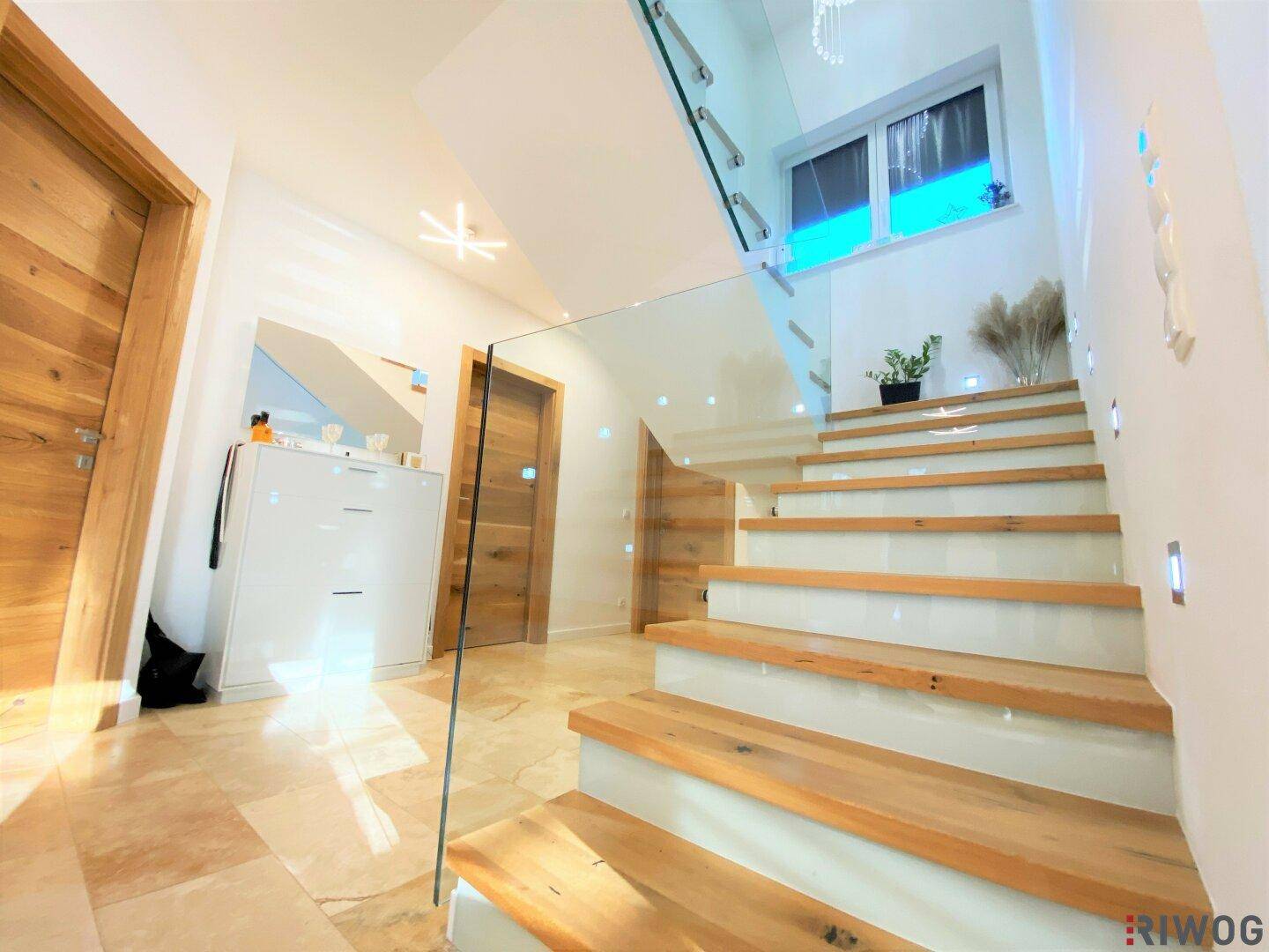 Moderner Eingangsbereich mit eleganter Holztreppe, Glasgeländer und hellem Fliesenboden.