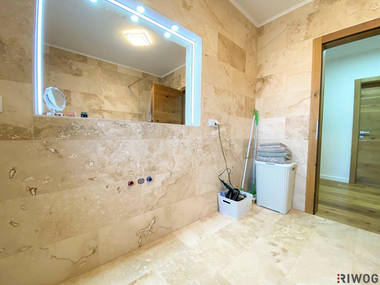 Badezimmer mit großem beleuchteten Spiegel und beigefarbenen Natursteinfliesen, bereit für die Einrichtung.