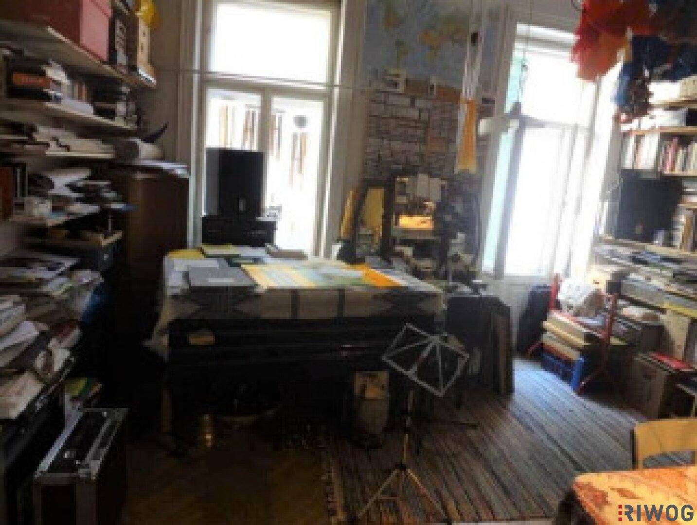 Überfüllter Raum mit hohen Fenstern, Holzboden und vielen Büchern und Gegenständen.