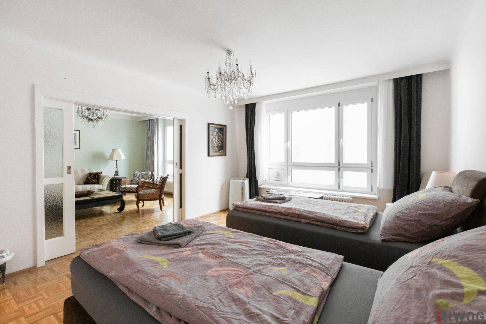Schlafzimmer mit zwei Betten, großen Fenstern und Blick in das angrenzende Wohnzimmer.