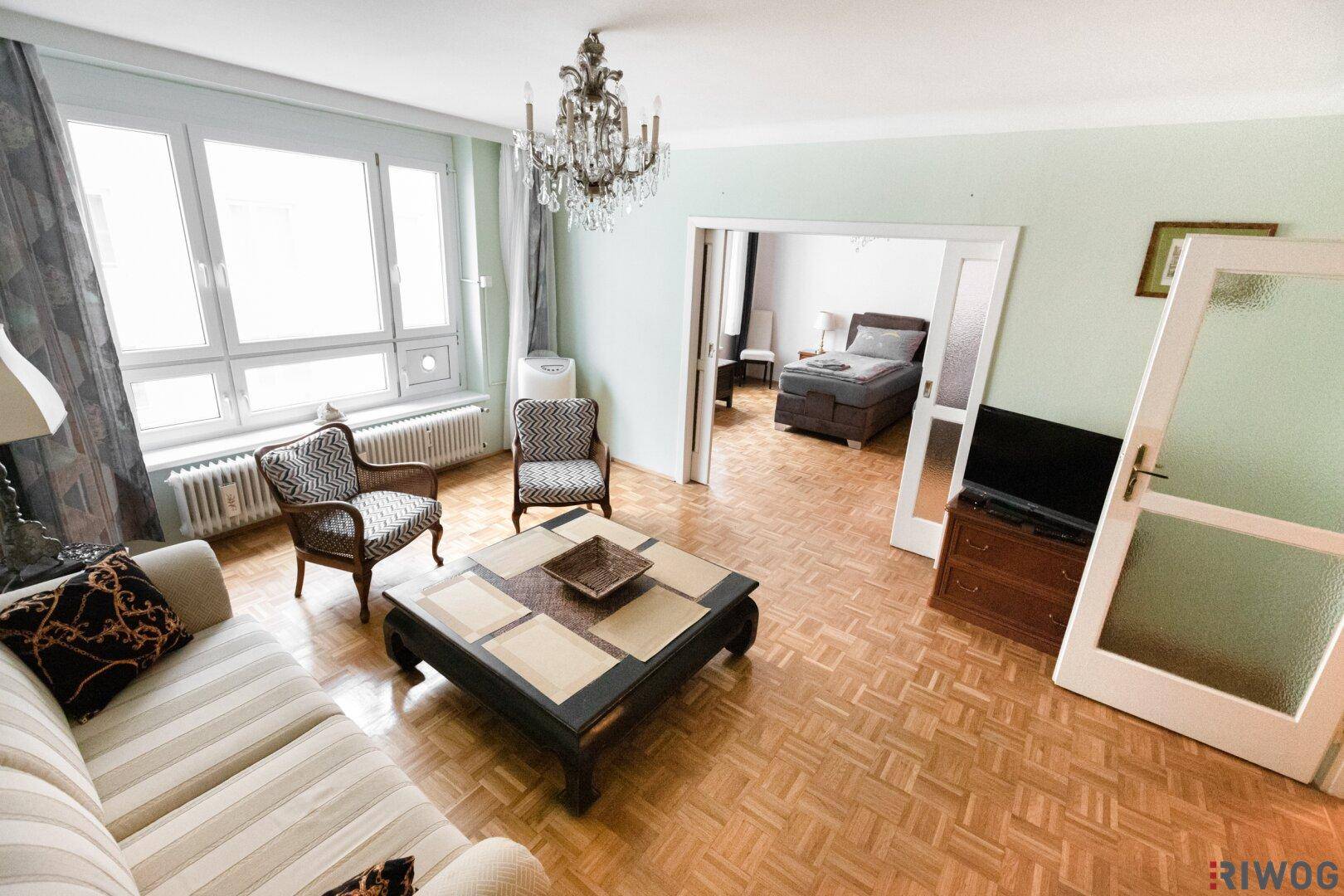 Helles Wohnzimmer mit Fischgrätparkett, Kronleuchter und Zugang zu einem weiteren Zimmer.