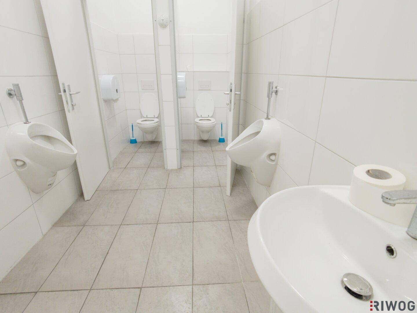 Sauberer Toilettenraum mit weißen Fliesen, mehreren Urinalen und Toilettenkabinen.