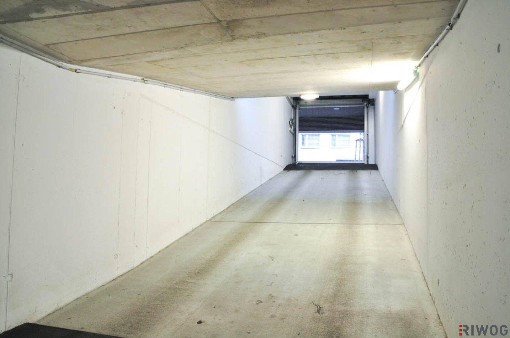 Einfahrt zur Tiefgarage mit Betonrampe und Rolltor, das Tageslicht hereinlässt.