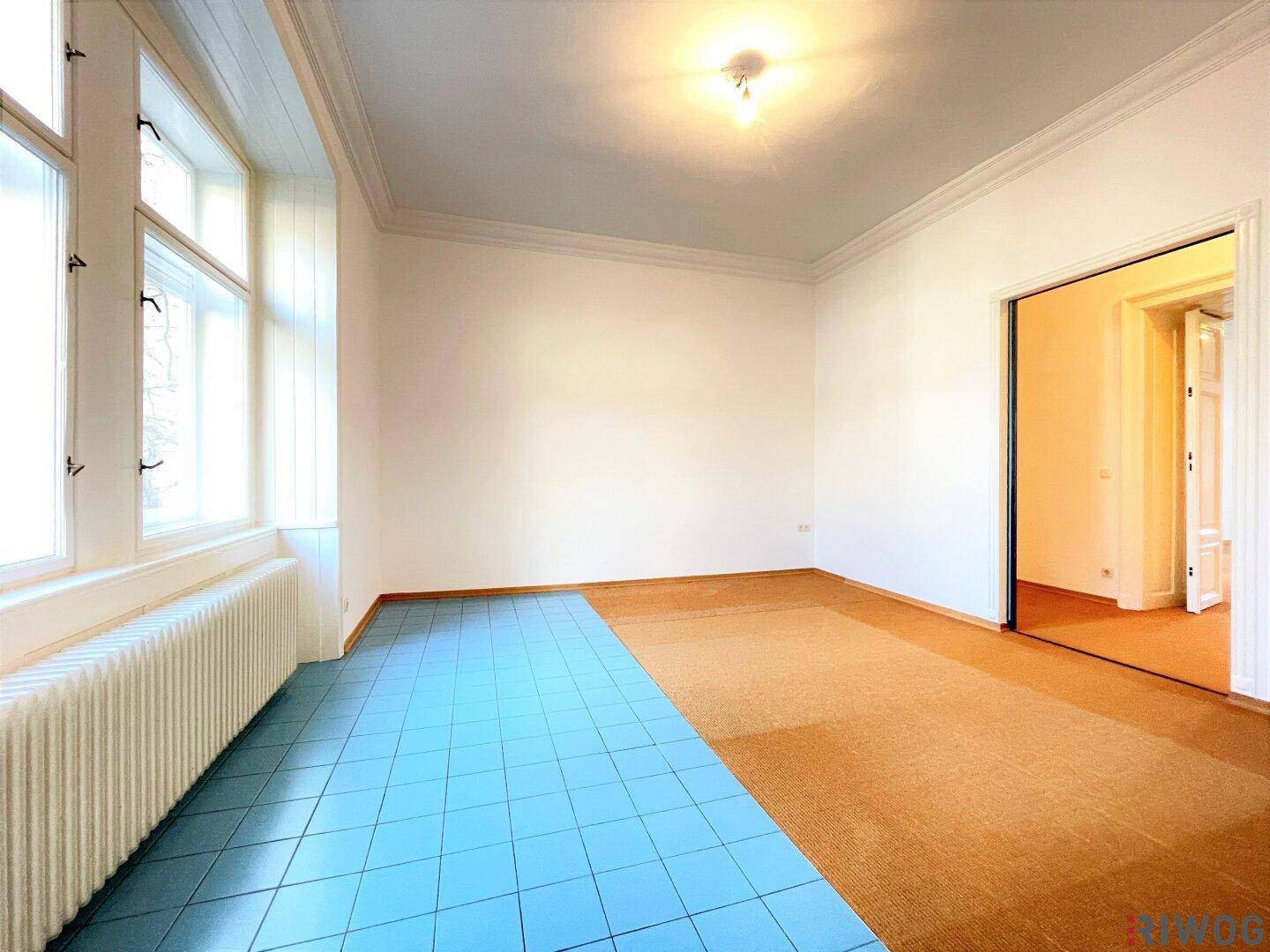 Zimmer mit blauen Fliesen und Teppichboden, hohen Fenstern und Heizkörper.