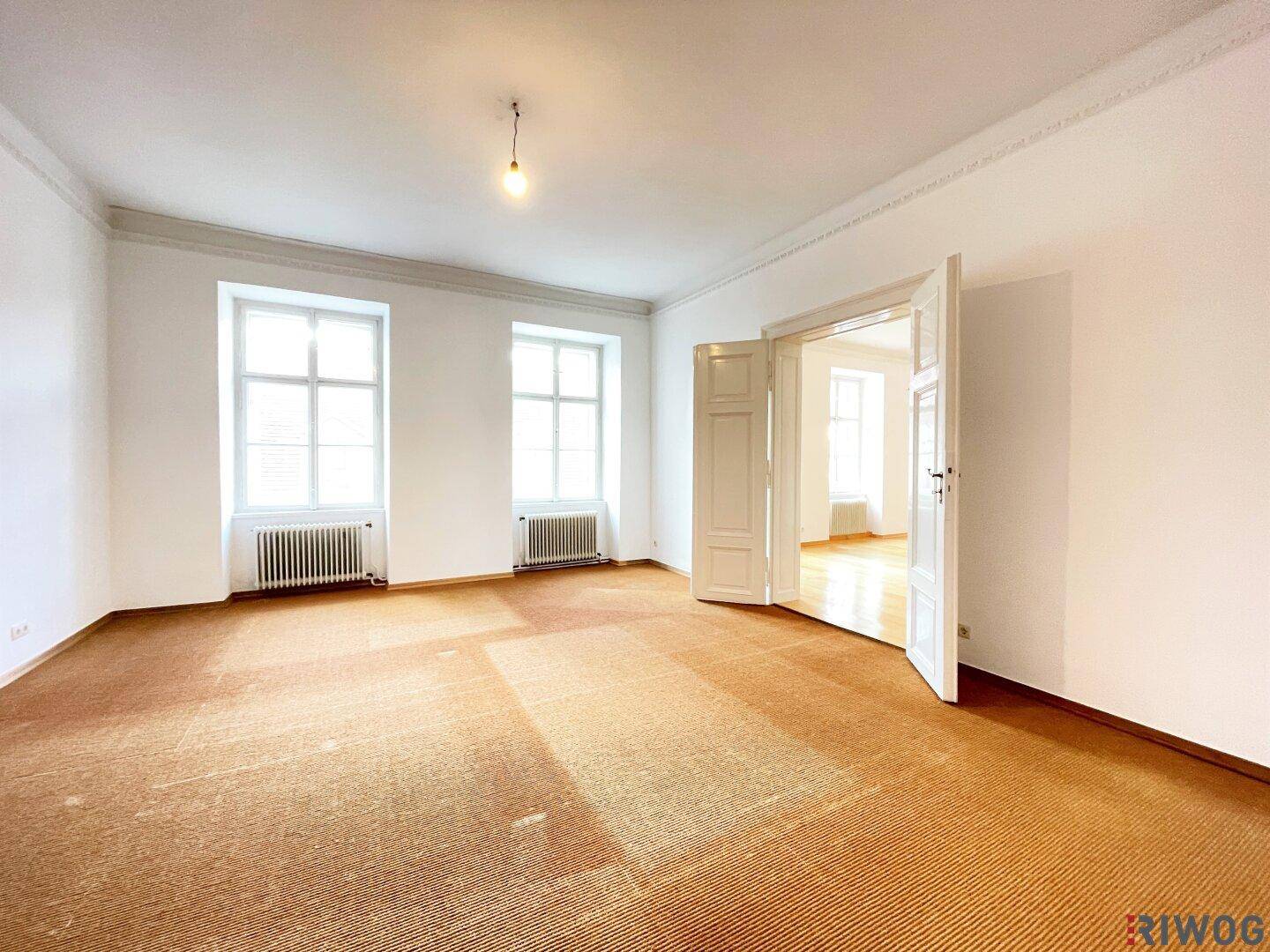 Geräumiges Zimmer mit Teppichboden, hohen Fenstern und Zugang zu einem weiteren Raum.