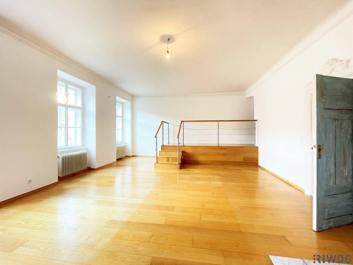 Geräumiges Wohnzimmer mit hellem Parkettboden, hohen Decken und einer Treppe zu einer oberen Ebene.